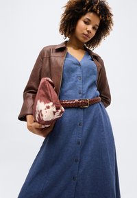 Γυναίκα με κατσαρά μαλλιά που φοράει μπλε φόρεμα με κουμπιά, καφέ πλεκτή ζώνη και καφέ δερμάτινο μπουφάν, κρατώντας μια τσάντα με μοτίβο σε μπορντό και λευκό χρώμα.