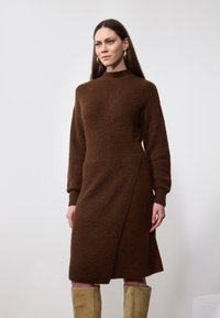 Robe tricotée marron à manches longues, col montant, design portefeuille et tissu texturé. Le mannequin porte des bottes beiges jusqu'aux genoux. Fond simple.