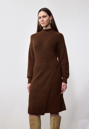 Brown knit dress with long sleeves, high neckline, wrap design, and textured fabric. Model wears beige knee-high boots. Simple background.