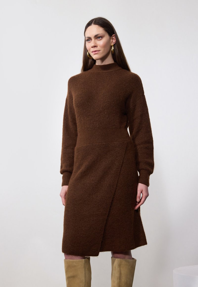 Robe tricotée marron à manches longues, col montant, design portefeuille et tissu texturé. Le mannequin porte des bottes beiges jusqu'aux genoux. Fond simple.