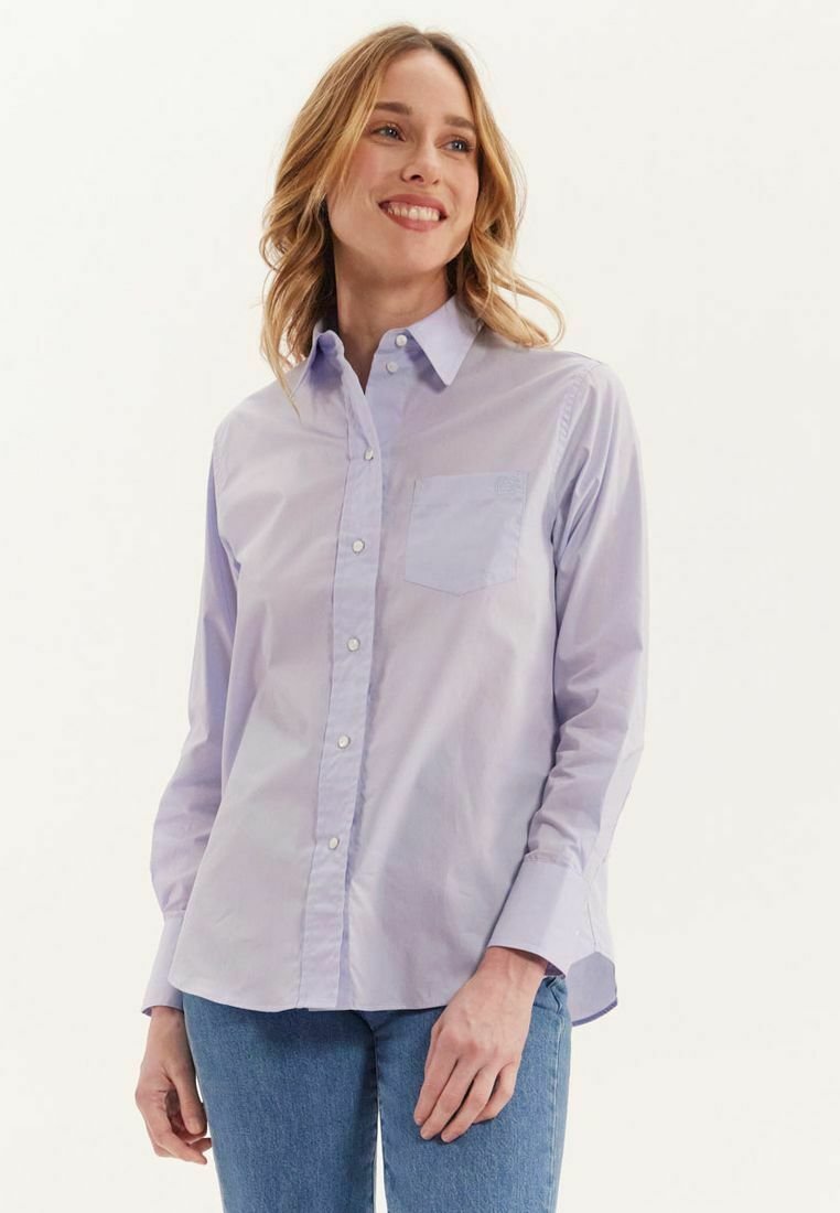 Chemise boutonnière mauve clair à manches longues, poche poitrine et texture lisse ; portée avec un jean bleu. Design simple et coupe décontractée.