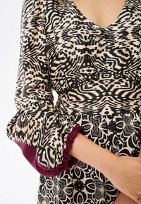 Zwart en crèmekleurige jurk met een V-hals, waarbij de volumineuze mouwen zijn versierd met bordeauxrode ruches en ingewikkelde zwarte bloemenpatronen.