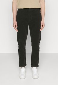 Pantalon noir en velours côtelé avec une coupe slim, doté d'une fermeture à boutons, de poignets roulés et de poches avant, associé à des baskets blanches.