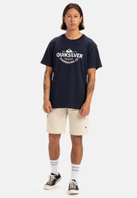 Camiseta de algodón azul marino con el logo blanco de Quiksilver, combinada con pantalones cortos beige, calcetines rayados blancos y zapatillas negras. Casual, con un ajuste relajado.