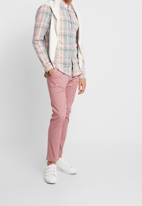 Hellblaue karierte Knopfhemd mit rosa, grünen und weißen Tönen, unter einem weißen Pullover getragen. Rosa schmal geschnittene Hose und weiße Sneakers.