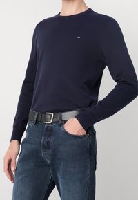 Pull à manches longues bleu marine en coton avec un col rond et un petit logo. Associé à un jean en denim foncé et une ceinture en cuir noir avec une boucle argentée.