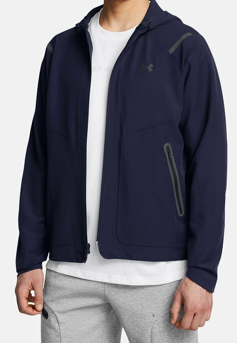 Homme portant une veste zippée bleu marine avec capuche, un t-shirt blanc et un pantalon de jogging gris clair, debout devant un fond uni.