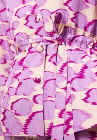 Veste en tissu à motifs violet et beige avec des boutons assortis et une ceinture nouée à la taille, montrant des détails de poche et de manches froncées.