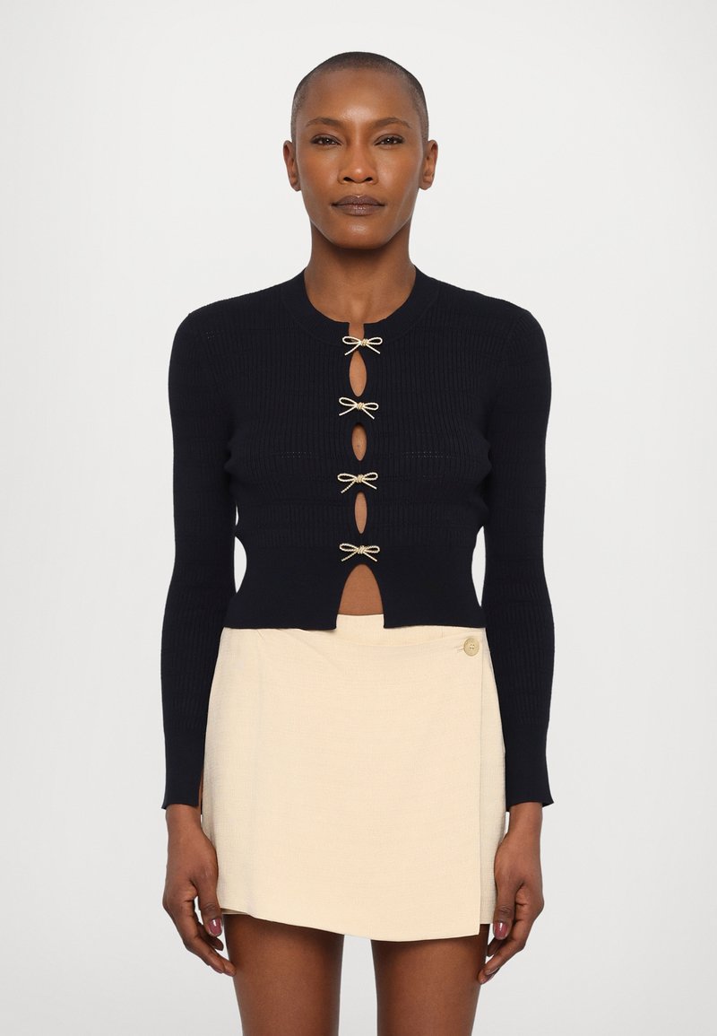 Mornarska rebrasta kardigana s okruglim izrezom, s prorezima ukrašenim bež mašnicama, u kombinaciji s krem mini suknjom s detaljem dugmeta.