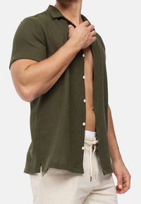 Chemise à manches courtes vert olive en tissu texturé, avec un col et des boutons blancs, partiellement déboutonnée, portée sur un pantalon de couleur claire.
