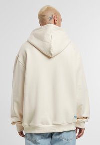 Béžová oversized mikina s velkou kapucí, žebrovanými manžetami a lemem, se malým modrým logem blízko spodního okraje.