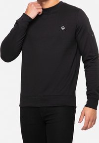 Sweat-shirt noir à manches longues, col rond, tissu texturé, poignets ajustés et petit logo en diamant sur la poitrine. Comprend une poche latérale.