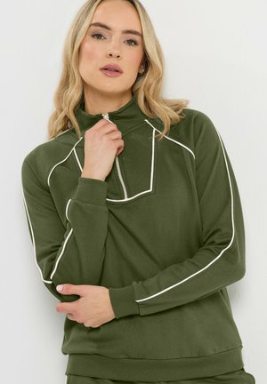 Femme aux cheveux blonds portant un sweat-shirt zippé vert avec des coutures blanches, posant avec une main près du menton et l'autre croisant son corps.