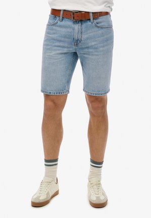 Pantaloni shorts in denim azzurro chiaro con gamba dritta, orlo sfrangiato e cintura in pelle marrone. Indossati con sneakers color crema e calze a righe.