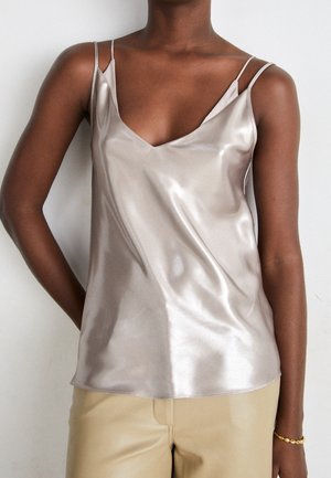 Dark-skinned person wearing a shiny silver satin camisole top with thin straps and beige pants against a plain light background.