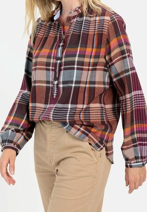 Femme portant une chemise à manches longues à carreaux multicolores avec col boutonné et pantalon beige, debout avec une main détendue le long du corps.
