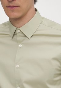 Uomo che indossa una camicia button-up beige chiaro con colletto, visibile dalla metà del petto fino sotto il naso, su sfondo semplice.