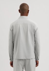 Lys grå jakke med jevn tekstur, lange ermer, krage og knappede mansjetter. Sett bakfra, viser ryddige sømmer og en avslappet passform.