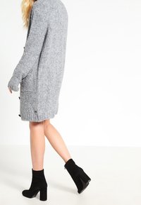 Grå stickad cardigan med en texturerad design, utrustad med knappar framtill. Kombinerad med svarta högklackade ankelboots.