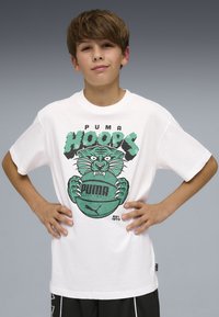 Αγόρι με κοντά καστανά μαλλιά, φοράει λευκό T-shirt Puma Hoops με πράσινη τίγρη που κρατάει μπάλα του μπάσκετ, στέκεται με τα χέρια στη μέση.