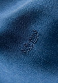 Denim-Stoff in tiefblau mit einem gestickten Logo-Akzent im Lorbeerdesign, das feine Nähte und eine glatte Textur zeigt.