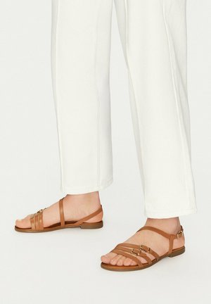 Stopy ubrane w białe spodnie i brązowe płaskie sandały z paskami i klamrami na białym tle.