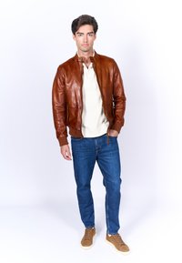 Bruin leren bomberjack met een ronde kraag, ritsluiting en zijzakken, gecombineerd met een crèmekleurig shirt, blauwe spijkerbroek en brown sneakers.