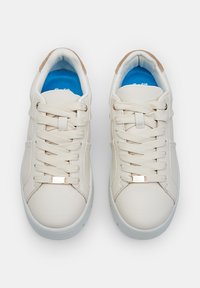 Baskets beige clair avec une tige synthétique lisse, des lacets plats, des semelles intérieures bleues et des accents métalliques subtils sur les boucles des lacets.