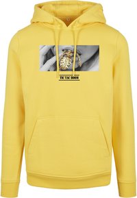 Žlutá mikina s kapucí s předním grafickým motivem, který zobrazuje černobílý obrázek ruky držící zlatý náhrdelník. Klokaní kapsa v přední části.