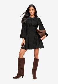 Musta tekstuuriga kleit, millel on puhvis varrukad, laienev seelik ja dekoratiivne vöökoht. Kombineeritud pruuni nubukist põlvepikkuste saapade ja käekotiga.