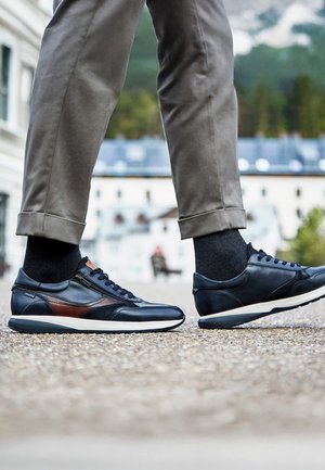 Beneden met grijze broek en zwarte sneakers met bruine accenten die op een grindpad staan in een stedelijke buitenomgeving.