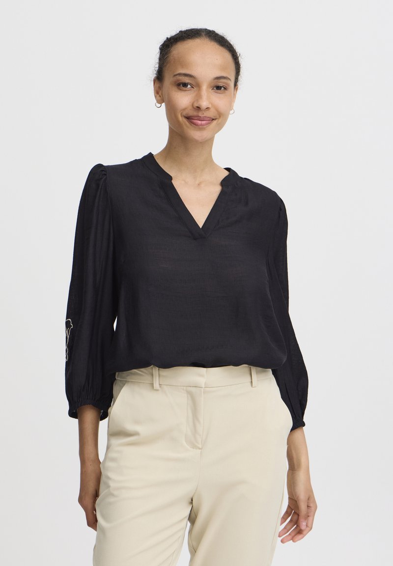Blusa nera a maniche lunghe con scollatura a V e spalle arricciate; presenta dettagli ricamati sui polsini; abbinata a pantaloni beige chiaro.