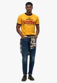 Yellow short-sleeve t-shirt with red "SuperMake" graphic, black trim. Denim jeans, plaid shirt tied at waist, and black shoes.