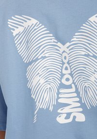 Lys blå t-shirt med et stort hvidt sommerfuglegraphic med fingeraftrykslignende linjer og ordet "SMILODON" i midten.