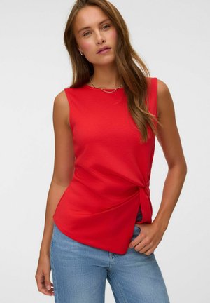 Femme aux longs cheveux bruns, portant un haut rouge sans manches noué à la taille et un jean bleu clair, debout devant un fond uni.