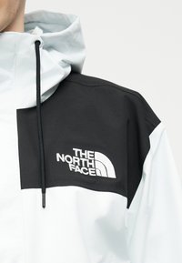 Mężczyzna ubrany w białą i czarną kurtkę z kapturem, z czarnym sznurkiem i naszytym na piersi logo "The North Face".