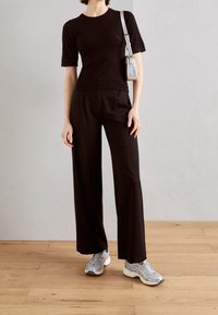 Person som bär en svart kortärmad topp och svarta vida byxor med grå sneakers och en liten silverfärgad axelväska.