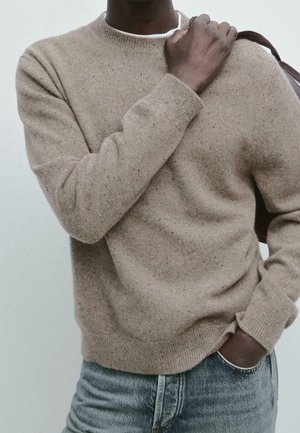 Mand i beige, plettet sweater med hånden, der holder en brun taskerem over skulderen, den anden hånd i lommen, iført lyseblå jeans.