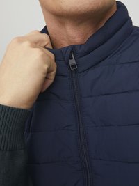 Gilet matelassé marine avec un col haut, fermeture éclair et motif matelassé. Présente un détail de manches en tricot côtelé et une texture externe lisse.