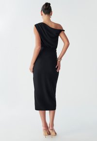 A woman stands in a fitted black dress, featuring an off-shoulder design, paired with beige heels. She has her hair tied up neatly.