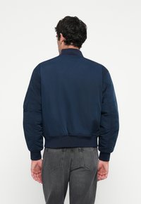 Man facing away wearing a navy blue bomber jacket and dark gray jeans against a plain white background.