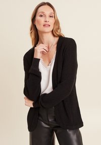 Cardigan noir en tricot texturé avec des manches longues, une ouverture à l'avant et des détails côtelés, porté par-dessus un haut en dentelle blanche et un pantalon noir.