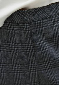 Tissu à carreaux gris avec un motif texturé, comportant des carrés et des accents à pied-de-poule. Détails de l'ourlet et de la taille visibles en haut.