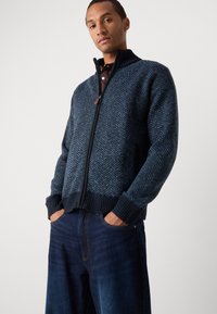 Pulover cu fermoar de culoare albastru închis, cu un model în zig-zag, guler și manșete ribbate, asortat cu blugi negri. Material tricotat texturat, două buzunare.