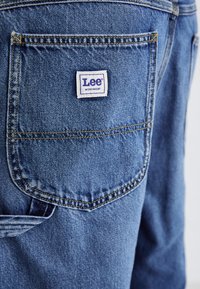 Denim työvaatteet farkut, joissa on takatasku, jossa on sininen tikkaus ja Lee-logo. Materiaali on keskipainoinen ja siinä on teksturoitu pinta.