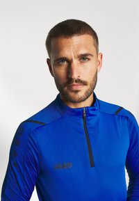 Blauwe sportieve longsleeve top met een kwart ritssluiting en kraag. Kenmerkt zich door gestructureerde schouderaccenten en een logo op de borst.