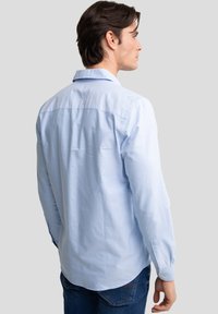 Mann, der nach rechts schaut, trägt ein hellblaues langärmeliges Hemd mit Knöpfen und dunkelblaue Jeans vor einem einfarbigen Hintergrund.