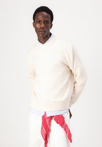 Pull en tricot couleur crème avec un col rond, porté par-dessus une chemise rayée bleu clair. Un bandana rouge est noué autour de la taille sur un pantalon blanc.