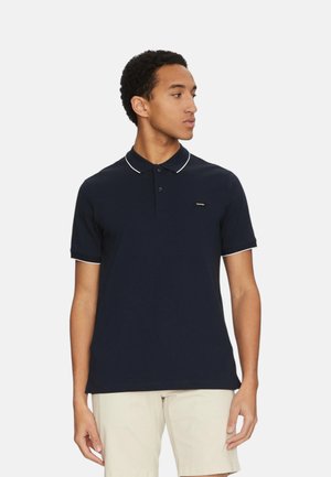 Uomo in piedi, guarda di lato, indossa una polo blu navy a maniche corte con profilo bianco e pantaloncini color beige chiaro su sfondo semplice.