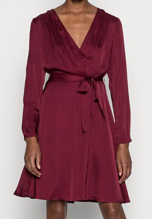 Žena v dlouhých rukávech, tmavě červených zavinovacích šatech s vázacím páskem v pase a výstřihem do V, stojící před jednobarevným pozadím.